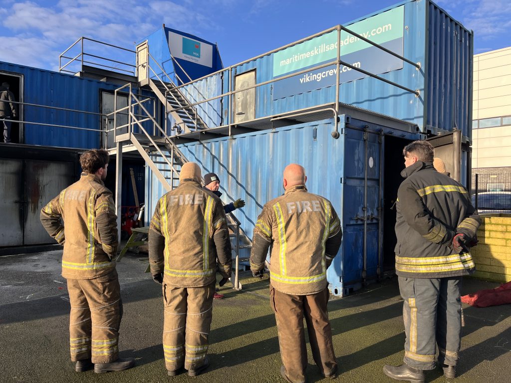 Delegates in fire fighting PPE with their backs to the camera, facing the instructor on the fire training ground | MSA Dover STCW Basic Safety Training Week