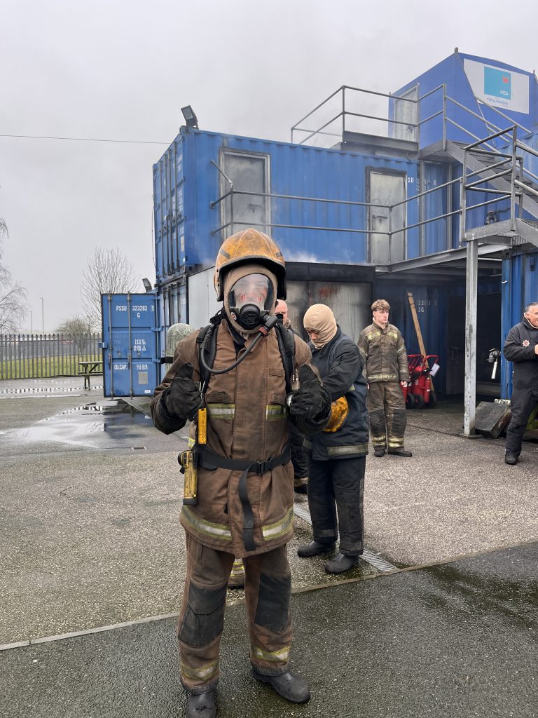 Finn stood in full fire fighting PPE with a helmet and breathing apparatus with his thumbs up to the camera as part of his 4-part STCW Basic Safety Training